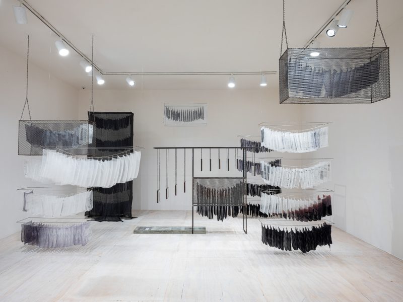 A white-wall gallery with legal documents printed on silk chiffon suspended from fishing line to resemble a police evidence room.