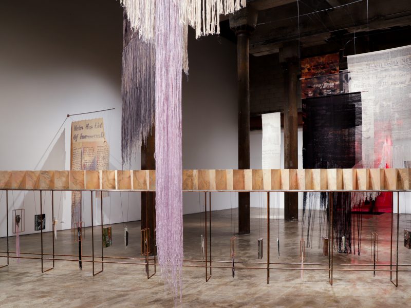 Purple threads hang in foreground, background is bisected by pages of accordion book spanning gallery.
