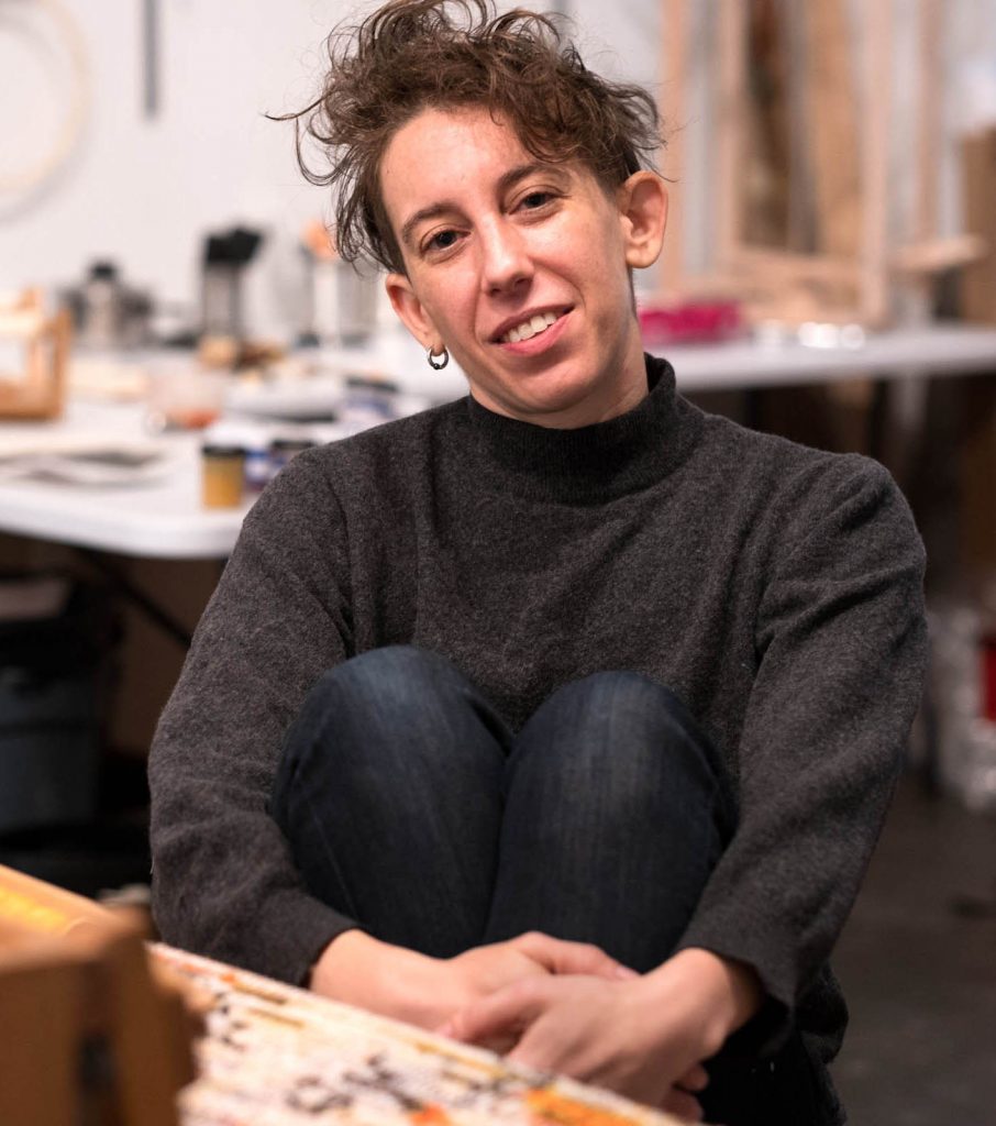 a white queer person sits in front of a loom, leaning back holding knees in hands and smiling