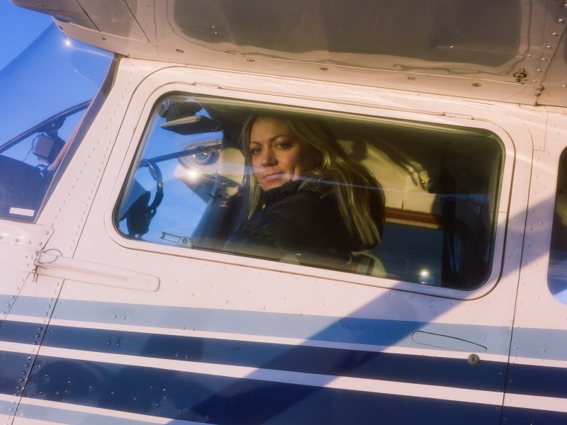 A white woman peers out of the cockpit of a small plane as the sun casts a pink glow.