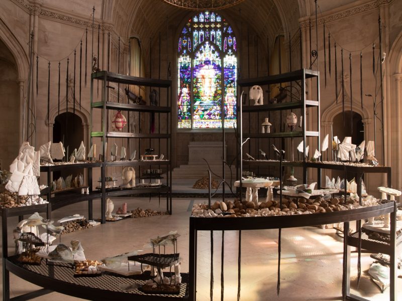 A metal circular sculpture inside a gothic-style chapel. On the circular shelves, which look like mausoleum crypts, there are carved marble sculptures, stones, fused glass pieces with archival fragments, and concrete urns. Around the circle are fabric tassels that look like fence posts.